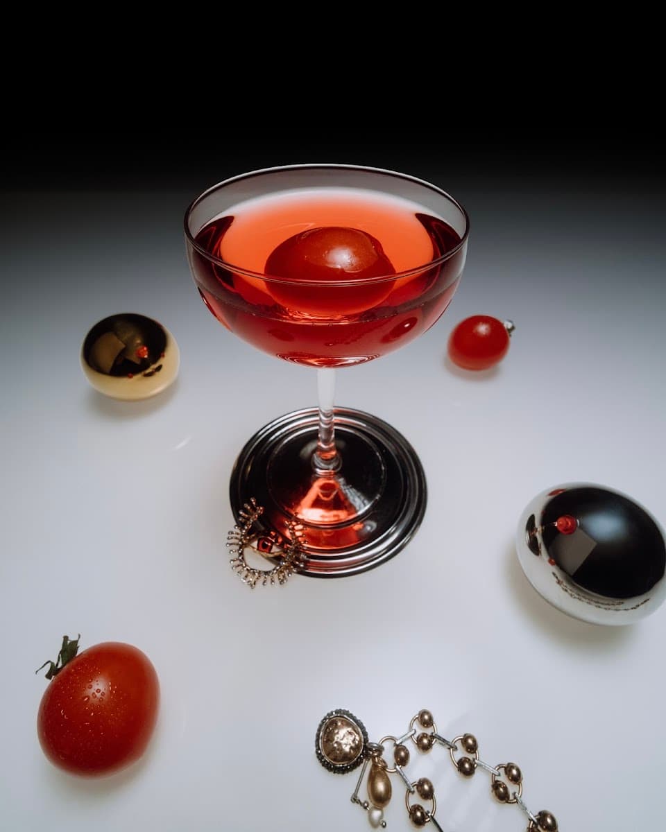 Pahar elegant cu cocktail roșu-portocaliu, garnisit cu o roșie cherry și accente aurii