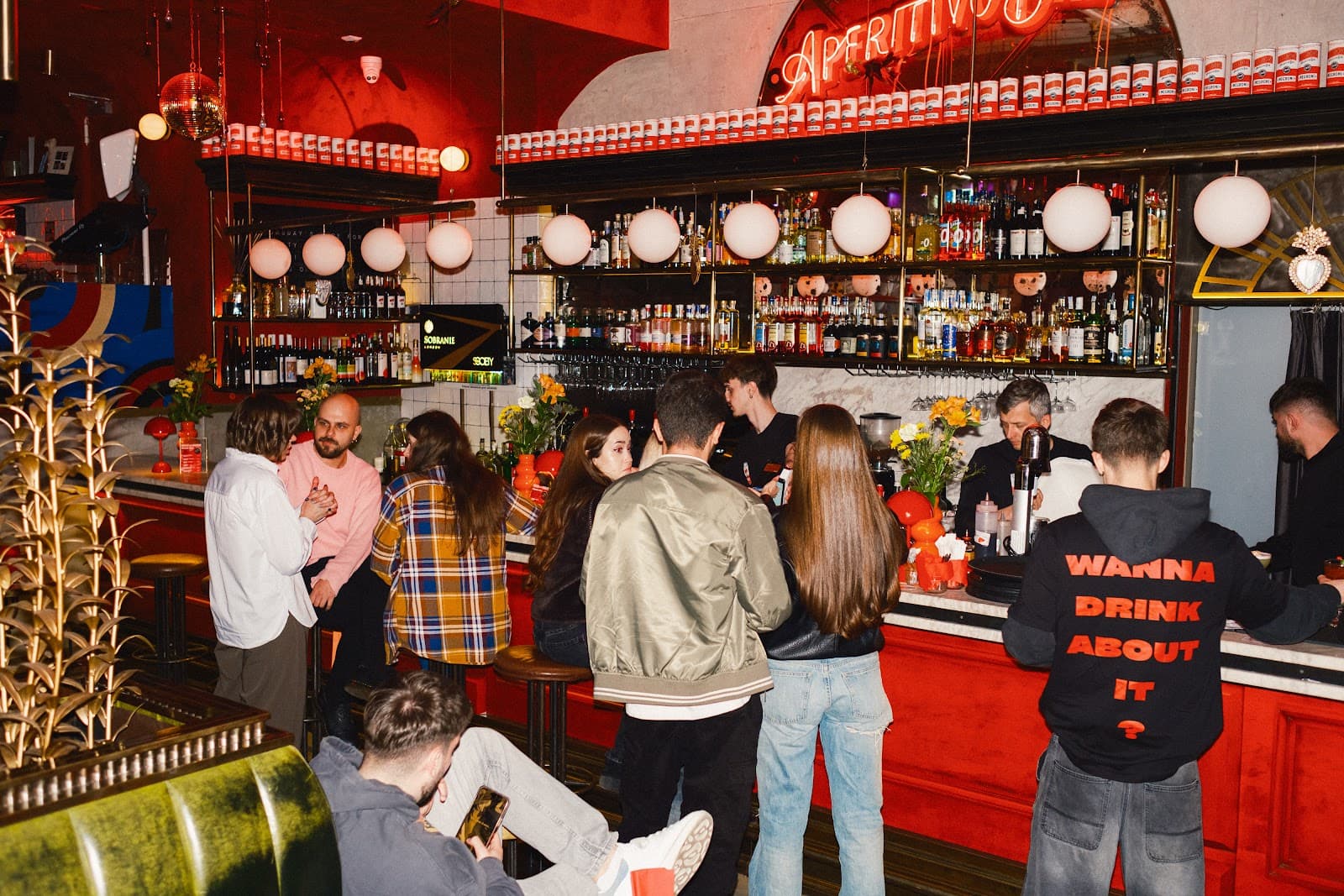Interiorul barului, cu oameni socializând la bar, decor roșu intens și rafturi pline cu sticle